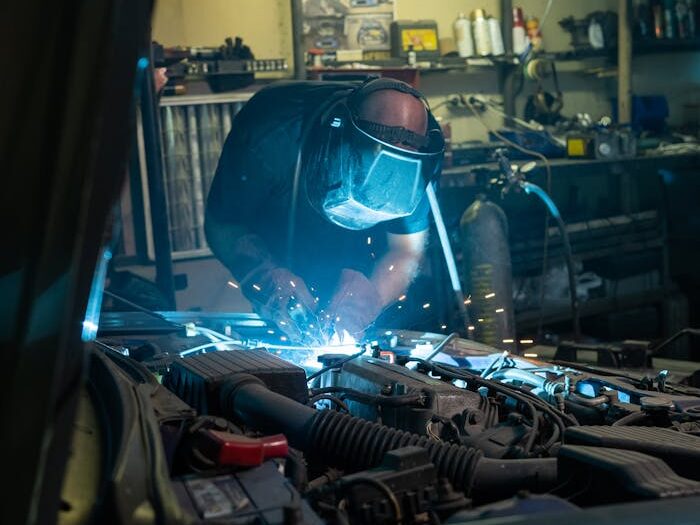 contact-img Mechanic wearing safety gear welding under hood of a car in a garage workshop.